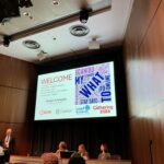 A conference room with a large screen displaying a welcome slide. Four people are at the front—one man stands at a podium, two women and a man are seated at a table. A word cloud is visible on the screen, along with organization logos.