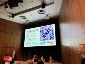 A conference room with a large screen displaying a welcome slide. Four people are at the front—one man stands at a podium, two women and a man are seated at a table. A word cloud is visible on the screen, along with organization logos.
